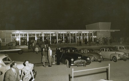 Black and white photograph of four cars parked in a row in front of a building. Speakers on a wire have been threaded through the driver’s front windows. There are also people standing around in semi-formal attire.